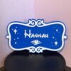 Front view of personalised cruise stateroom door plaque — custom name "Hannah" in white raised lettering on blue background with sparkle stars, scroll frame and winged charm, UK made 3D print