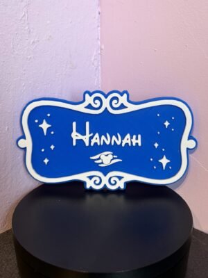 Front view of personalised cruise stateroom door plaque — custom name "Hannah" in white raised lettering on blue background with sparkle stars, scroll frame and winged charm, UK made 3D print