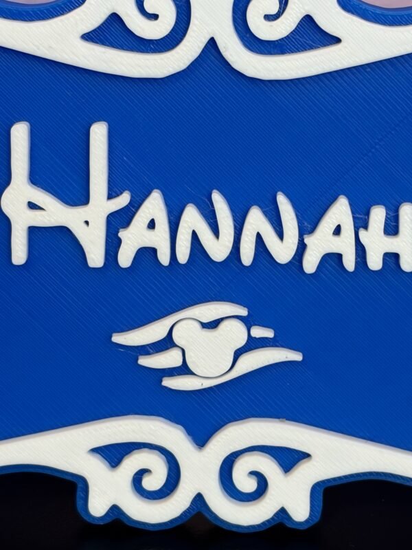 Close-up of personalised magical cruise stateroom door plaque showing "Hannah" in enchanted storybook font — blue and white 3D printed with ornate scroll detail and winged charm motif