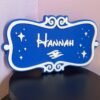 Side angle of personalised magical cruise door plaque — blue and white dual-colour 3D printed stateroom name sign with ornate scroll border and enchanted winged motif, freestanding display