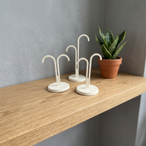 3D printed earring display stand set — three white minimal hook stands in tall asymmetric, short asymmetric and symmetric styles on round bases, styled on wooden shelf with snake plant, UK made jewellery display